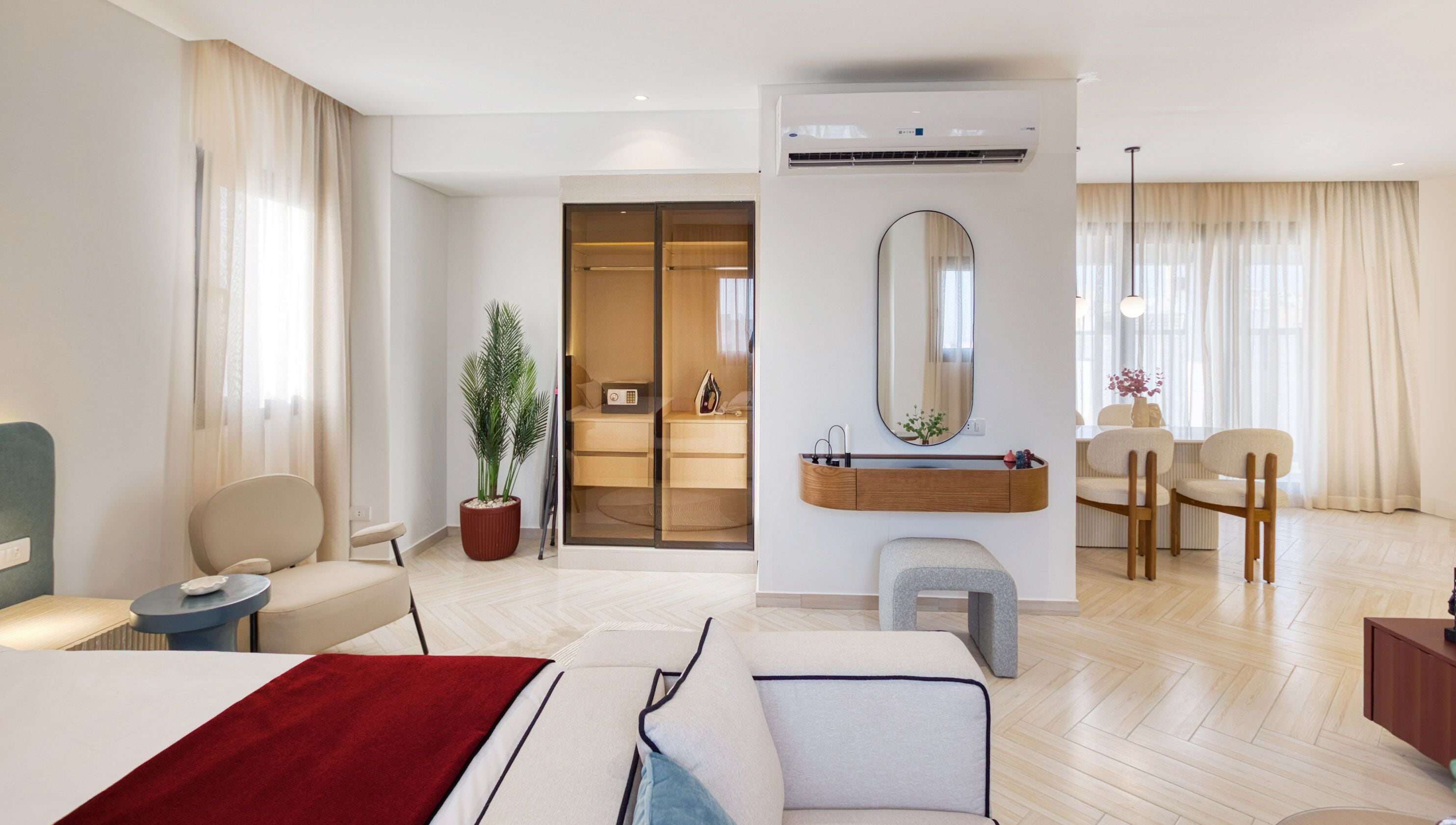 A modern hotel room with a bed, a desk, a chair, a small table, a potted plant, a mirror, and a built-in air conditioning unit.