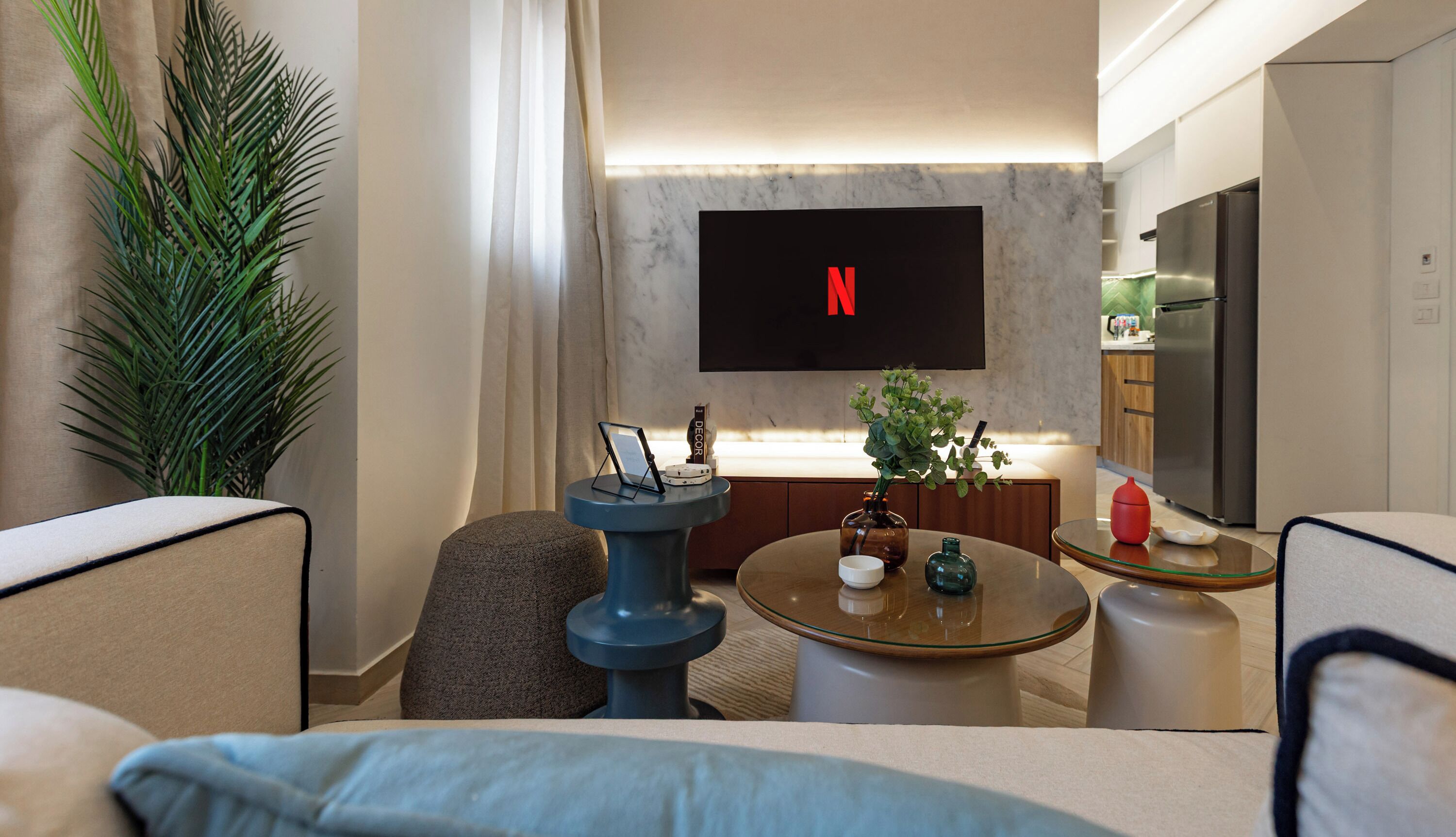 A modern living room with a flat-screen TV mounted on the wall, a coffee table, seating area, and a kitchen in the background.