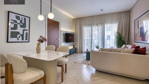 A modern living room with a sofa, a dining area with chairs and a table, a TV mounted on the wall, and a large window with curtains. - Prime Residence Zayed Hills (Sheikh Zayed City)