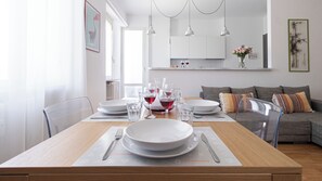 A dining area with a wooden table set for four, white plates, glasses, and cutlery. A gray sofa and a colorful cushion are in the background. - Italianway Comfort - Maffei 20 (Milan)