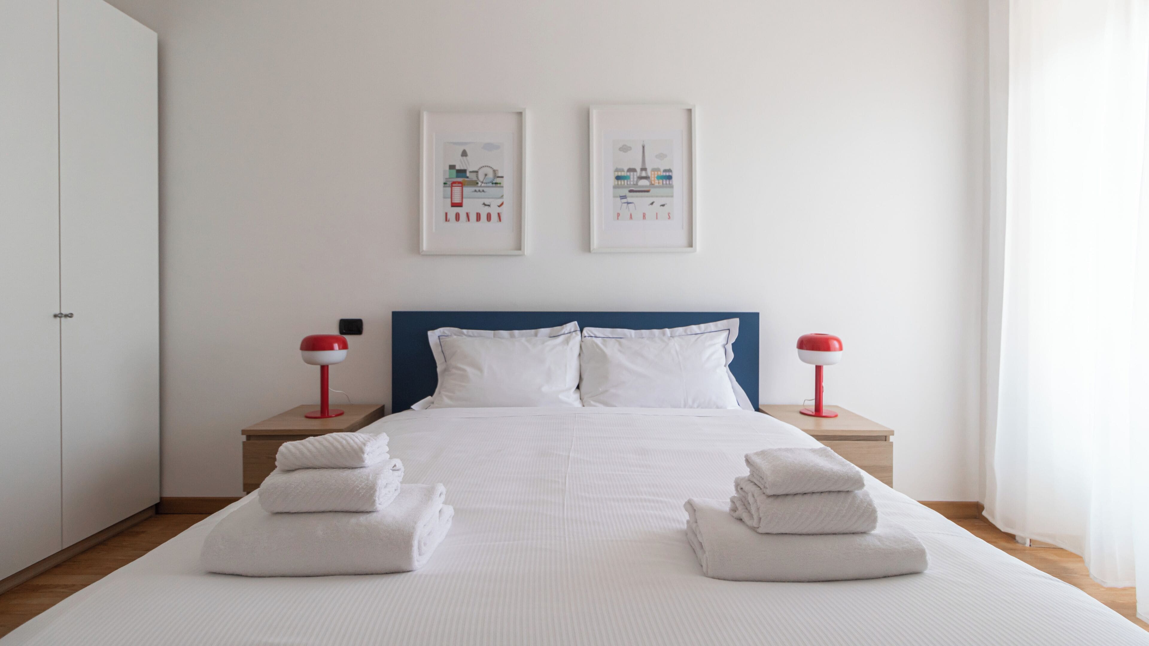 A neatly made bed with white linens and two red bedside lamps. There are two framed pictures on the wall above the bed, and towels are folded on the bed.