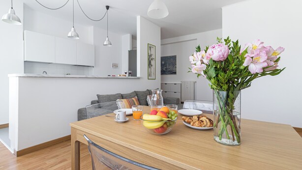 Una cocina moderna con armarios blancos, una mesa de comedor con fruta y pasteles, y un jarrón de flores.