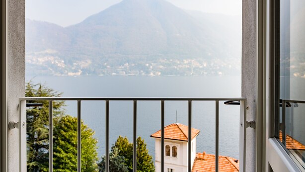 Un balcon doté d’une balustrade en métal, donnant sur un lac, avec une montagne en arrière-plan et un bâtiment surmonté d’un toit rouge.