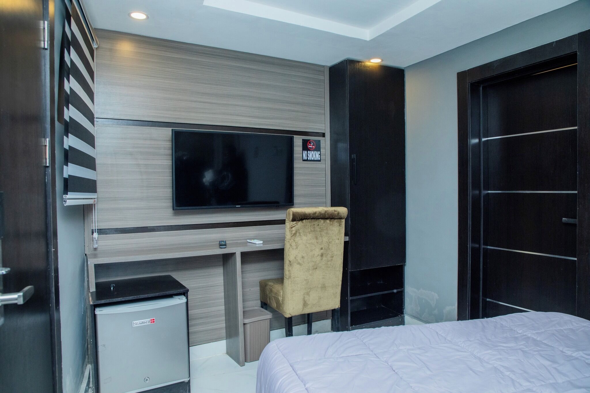 A modern hotel room with a bed, a desk with a television, a chair, and a wardrobe.