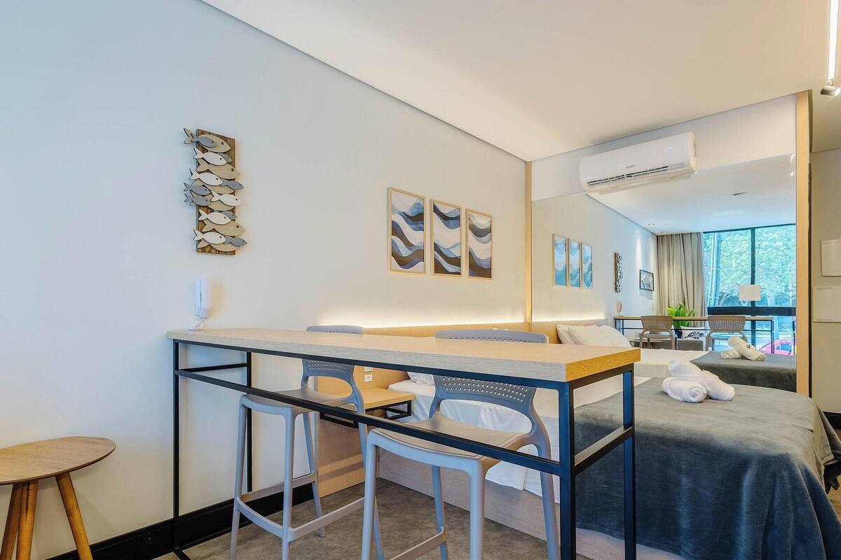 A modern hotel room with a bed, a desk with chairs, a small round table, artwork on the walls, and a large window.