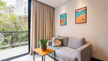 Ein modernes Wohnzimmer mit einem grauen Sofa, einem kleinen Holztisch mit einer Pflanze und einem großen Fenster mit Vorhängen.