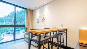 A modern dining area with a large table and chairs, a sofa, and a view of greenery through large windows.
