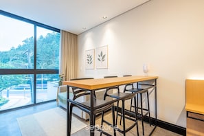 Un comedor moderno con una mesa grande y sillas, un sofá, y vistas a la vegetación a través de amplias ventanas.