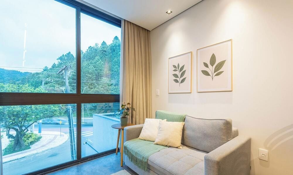 Een kamer met een groot raam, een bank en twee ingelijste bladerwerken aan de muur.
