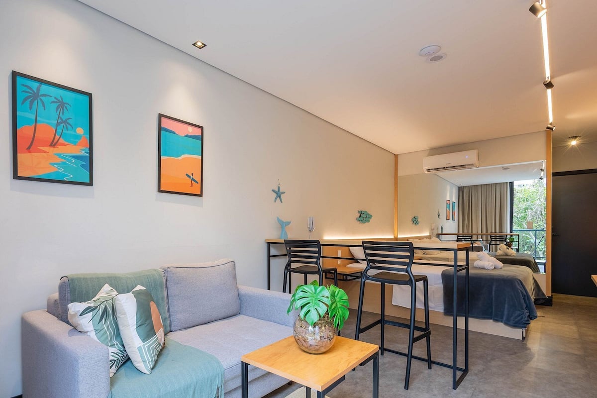 A modern hotel room with a sofa, a dining table, and a bed. There are two paintings on the wall, a starfish decoration, and a potted plant on the table.