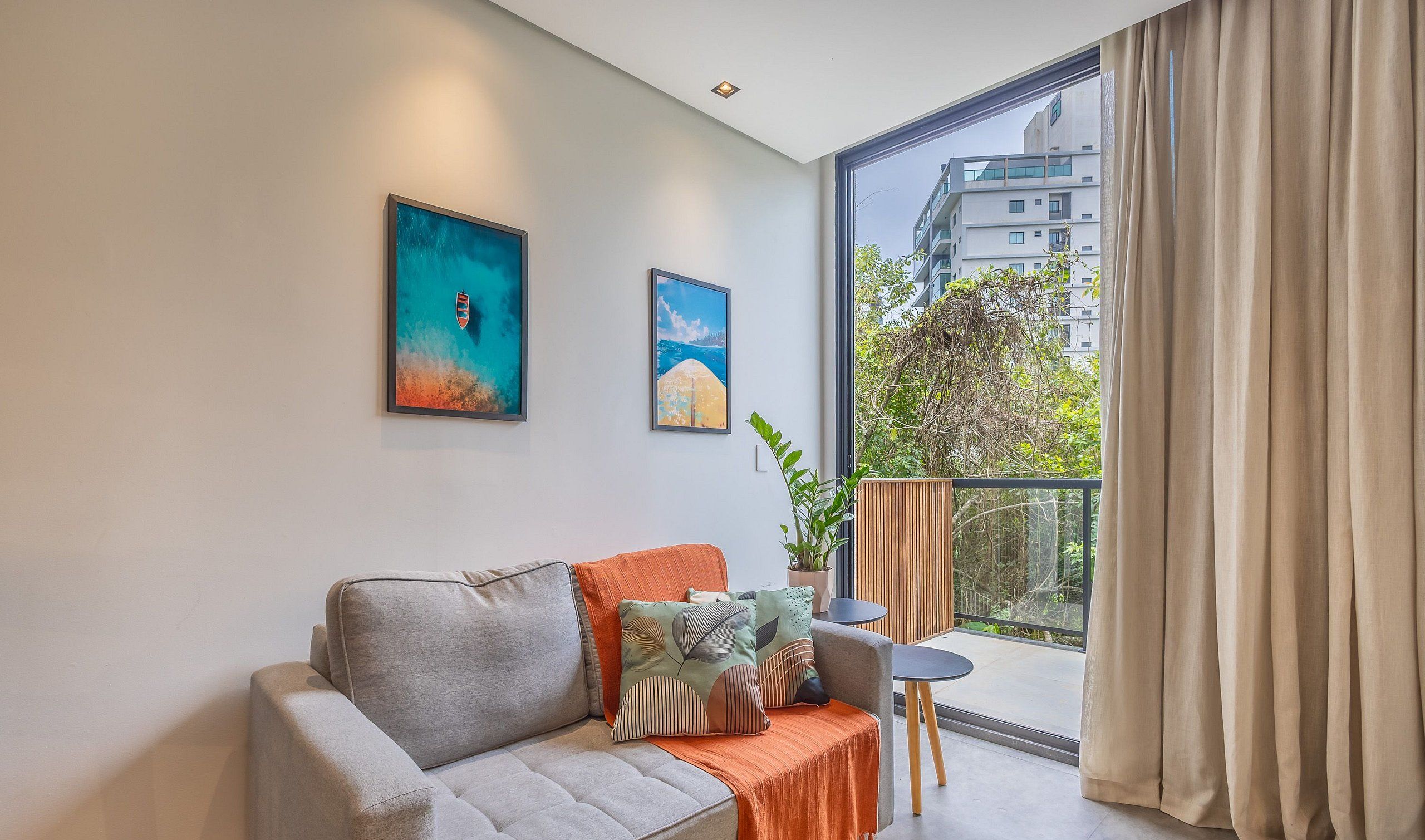 A modern living room with a grey sofa, orange throw, and two framed artworks. A small table and potted plant are on a balcony with a view of buildings and trees.