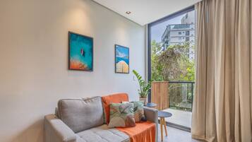 A modern living room with a grey sofa, orange throw, and two framed artworks. A small table and potted plant are on a balcony with a view of buildings and trees.