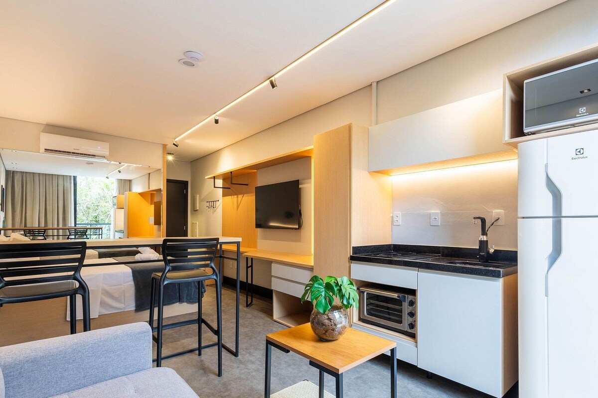 A modern hotel room with a kitchenette, a dining area with a table and chairs, a sofa, and a potted plant.