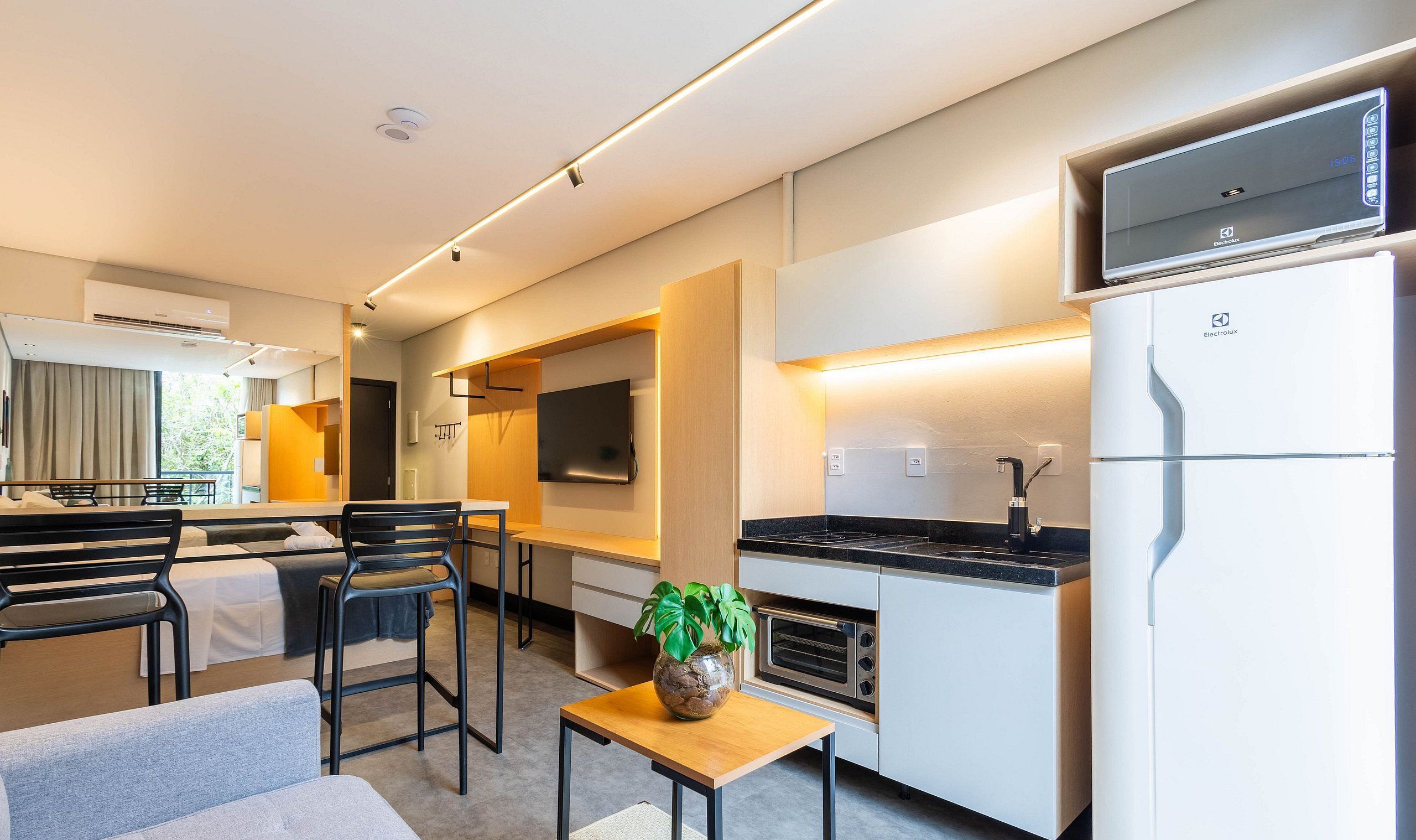 A modern hotel room with a kitchenette, a dining area with a table and chairs, a sofa, and a potted plant.