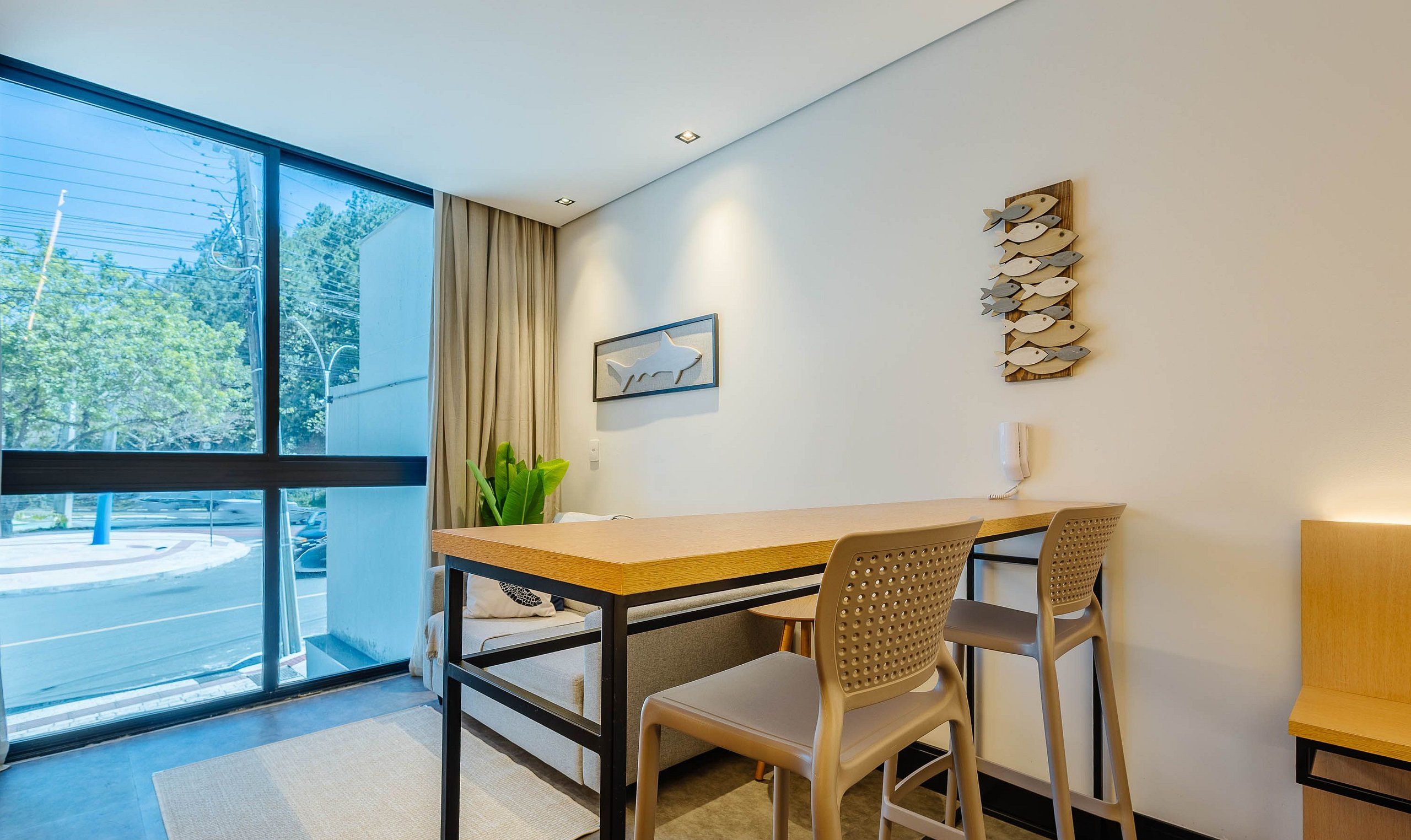 A modern room with a large window, a wooden table, two chairs, a potted plant, and a wall-mounted artwork.