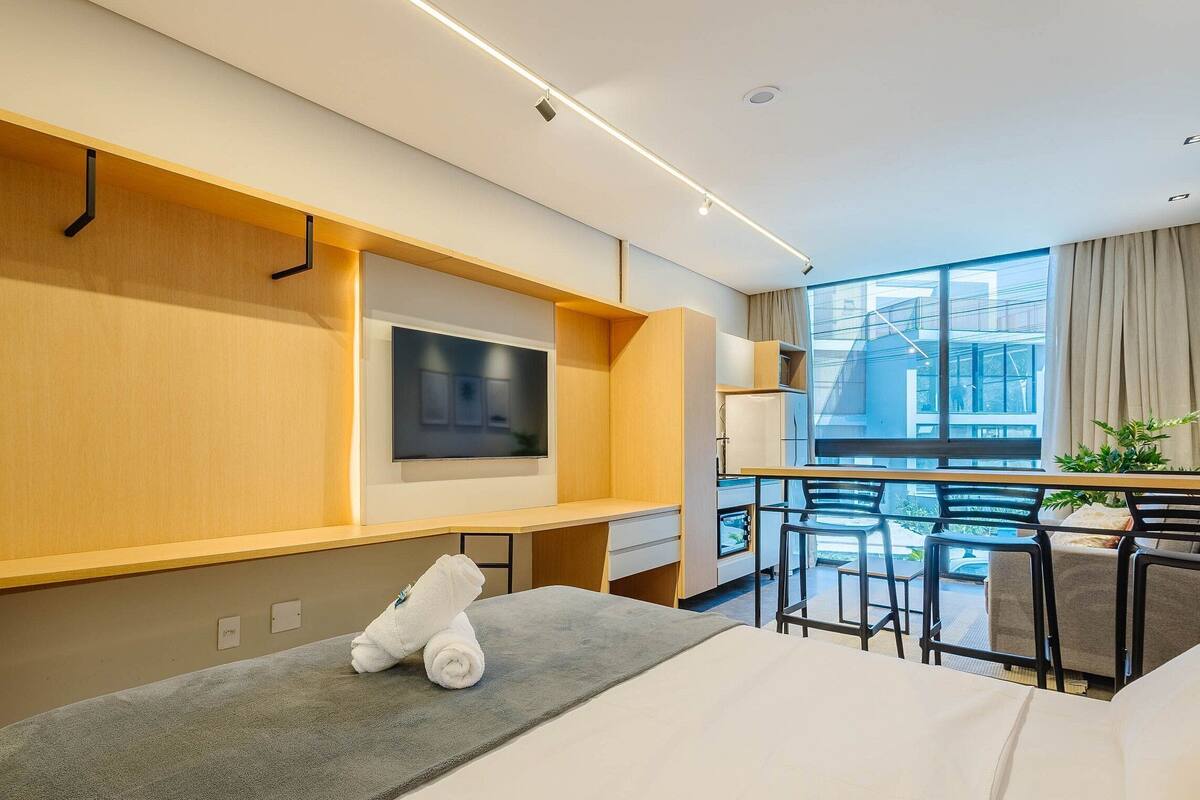 A modern hotel room with a large window, a flat-screen TV, a small desk, and a comfortable seating area.