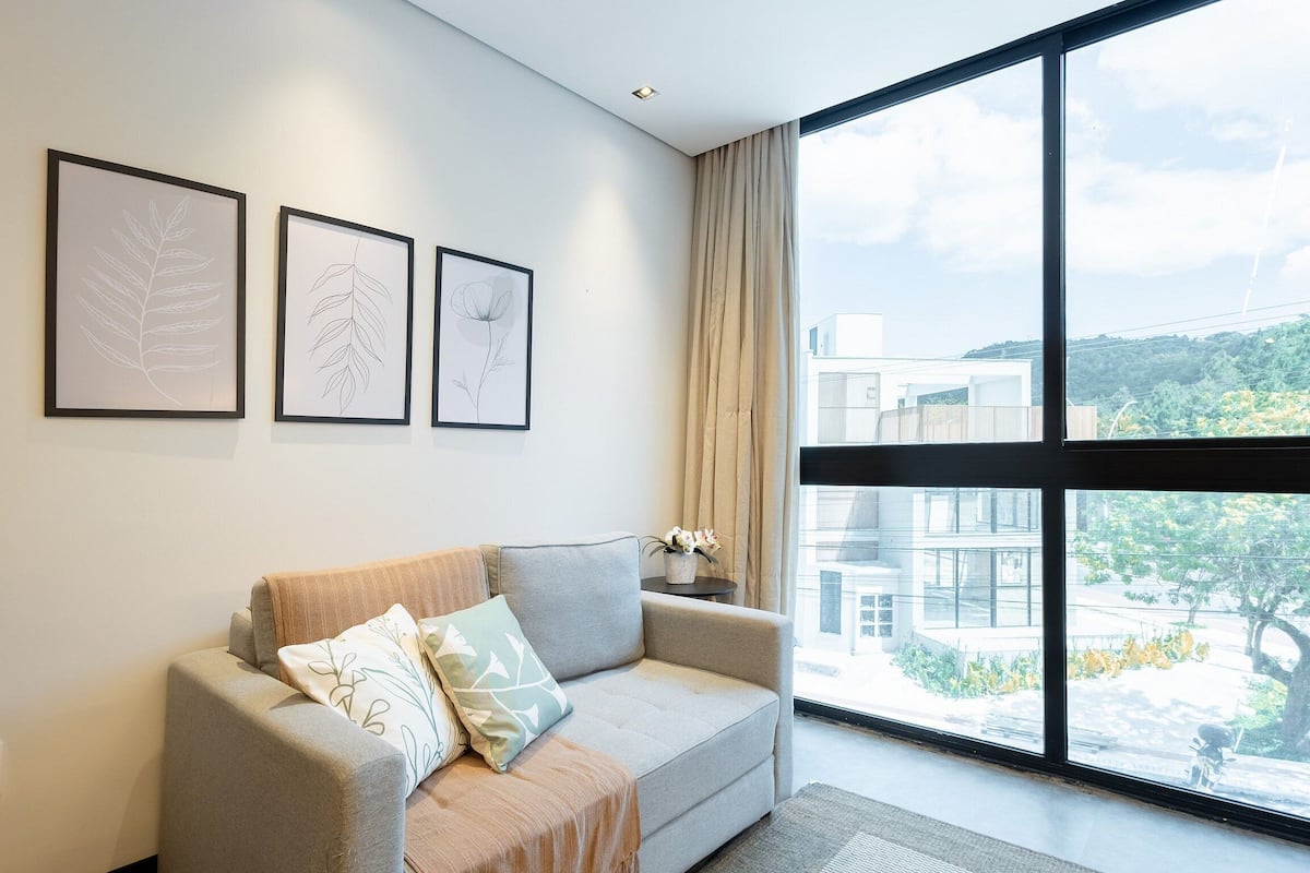 A modern living room with a grey sofa, three framed botanical prints on the wall, and a large window offering a view of buildings and greenery.