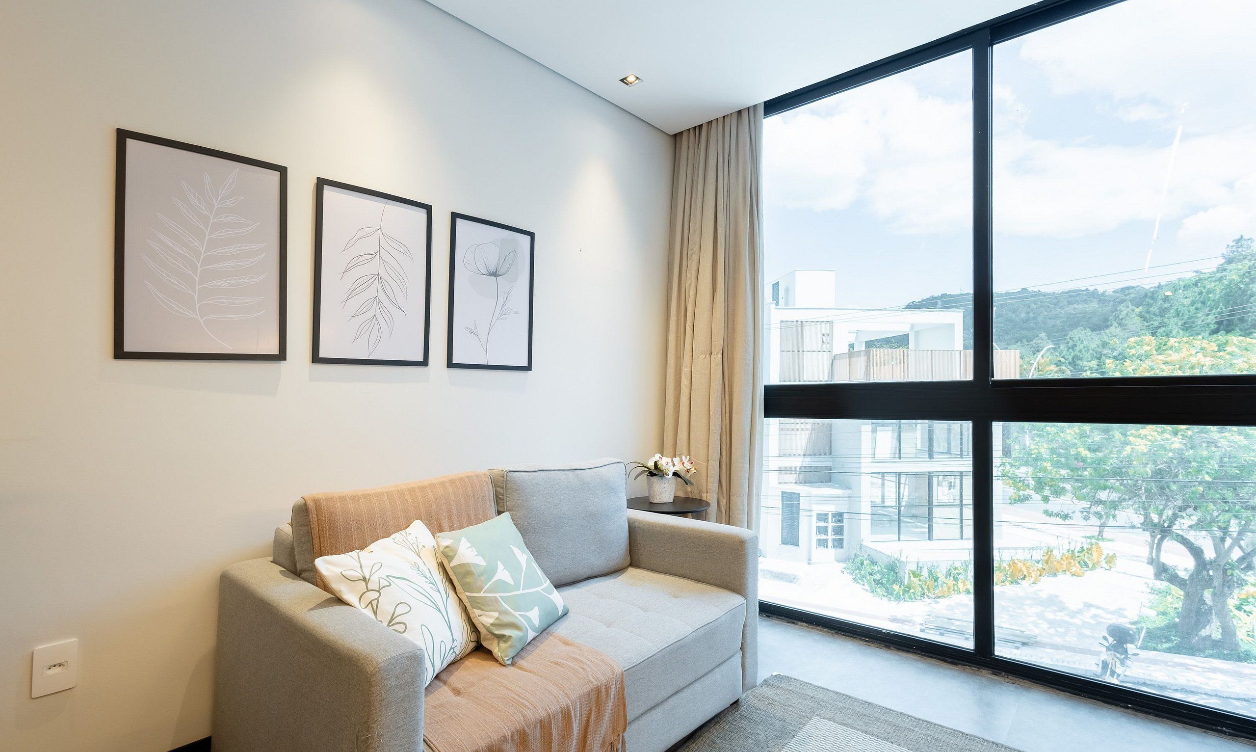 A modern living room with a grey sofa, three framed botanical prints on the wall, and a large window offering a view of buildings and greenery.