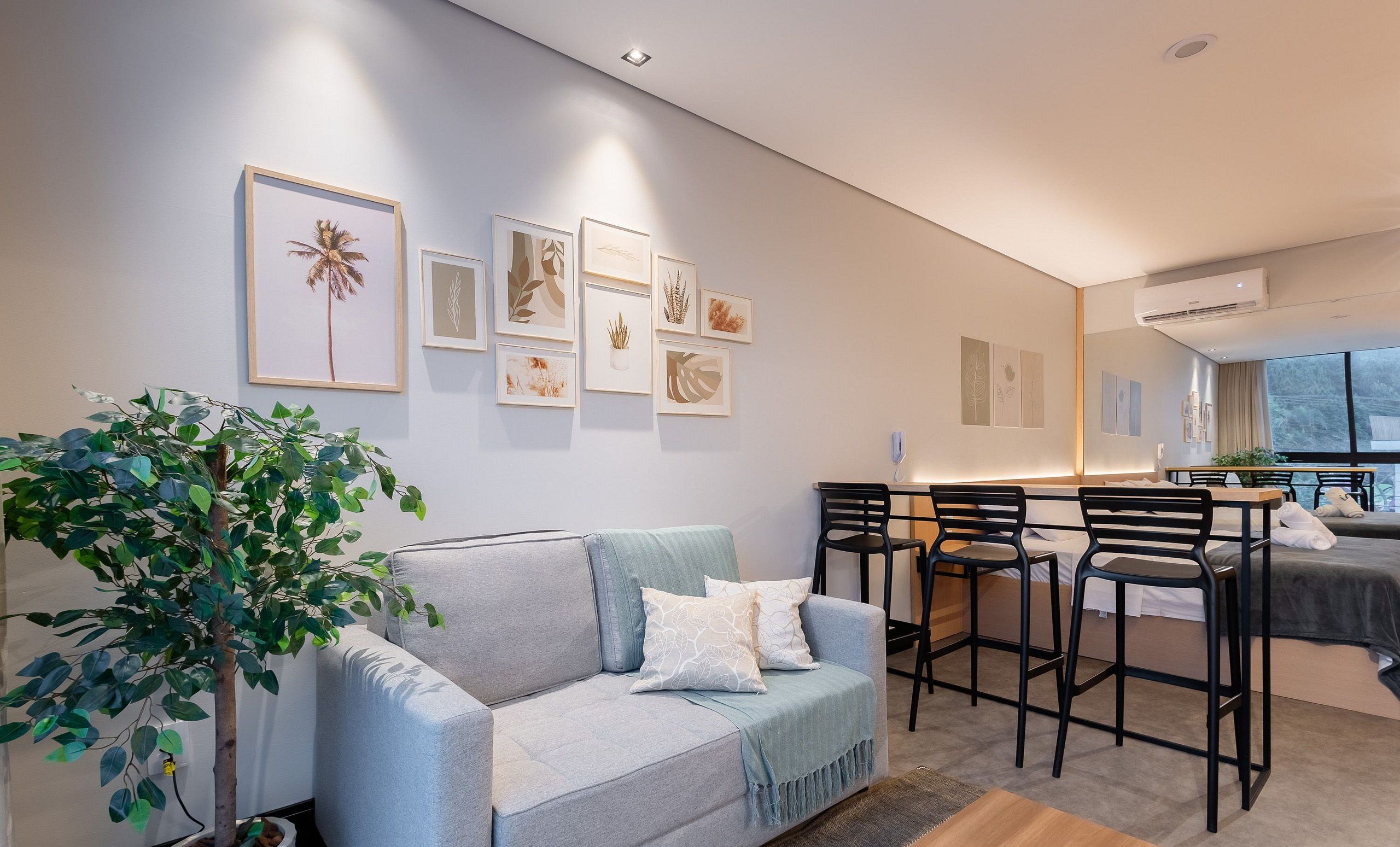 Een moderne woonkamer met een grijze bank, een salontafel en een eethoek met zwarte stoelen. Aan de muur hangen botanische prints in lijsten, en een groot raam zorgt voor natuurlijk licht.