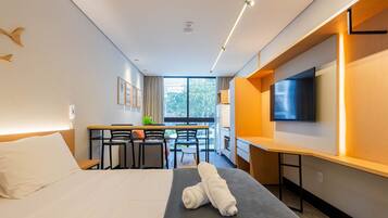 A hotel room with a bed, a desk with chairs, a TV, and a window with a view of the outside.