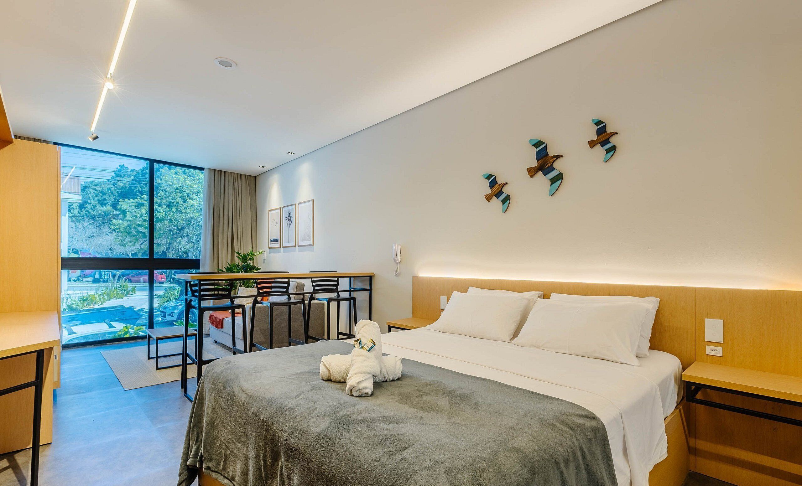 Een moderne hotelkamer met een bed, nachtkastjes, een bureau met stoelen, een groot raam en kunst aan de muur.