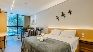 Une chambre d’hôtel moderne, équipée d’un lit, de tables de chevet, d’un bureau avec des chaises, d’une grande fenêtre et d’œuvres d’art accrochées au mur.