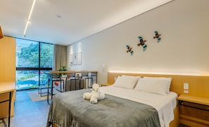 Une chambre d’hôtel moderne, équipée d’un lit, de tables de chevet, d’un bureau avec des chaises, d’une grande fenêtre et d’œuvres d’art accrochées au mur.