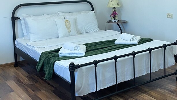 A single bed with a metal frame, white bedding, and a green throw blanket. There is a small bedside table with a lamp and a decorative item. The room has wooden flooring and white walls. - Warm House (Tbilisi)