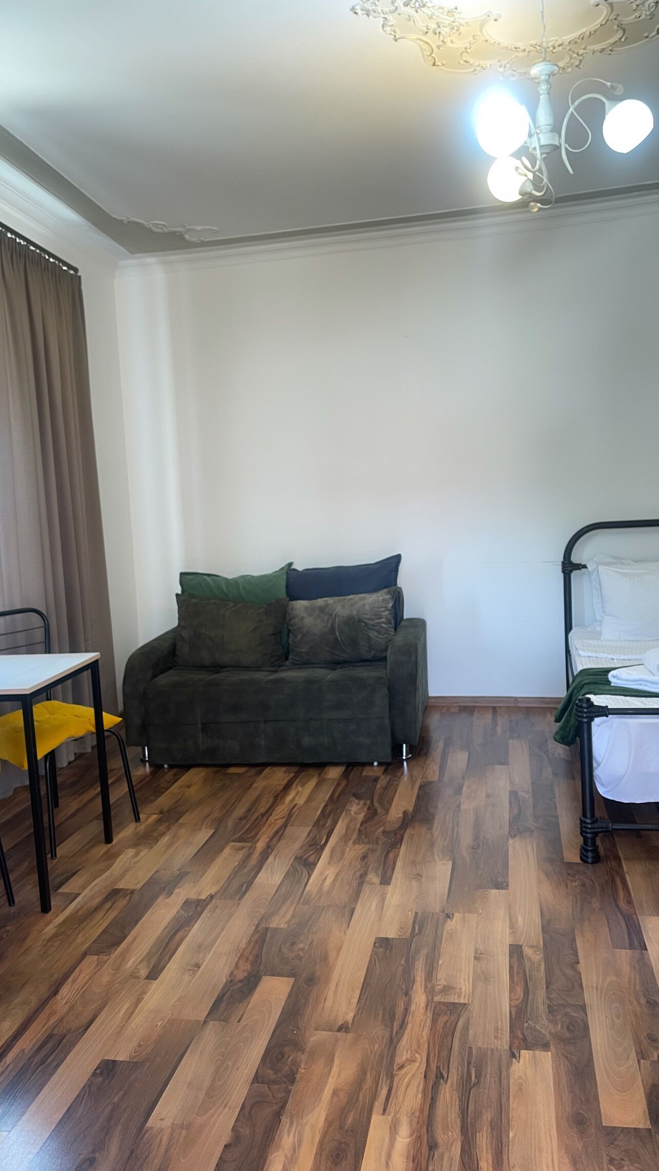 A room with a dark green sofa, a yellow chair, a bed with white bedding, and a wooden floor.