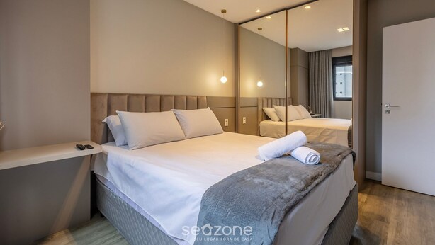 A modern hotel room with a large bed, a bedside table, a mirror, and a view of the outside. - IFT - Ed. Infinity Tower Flat 350m From Meia Praia (Itapema)