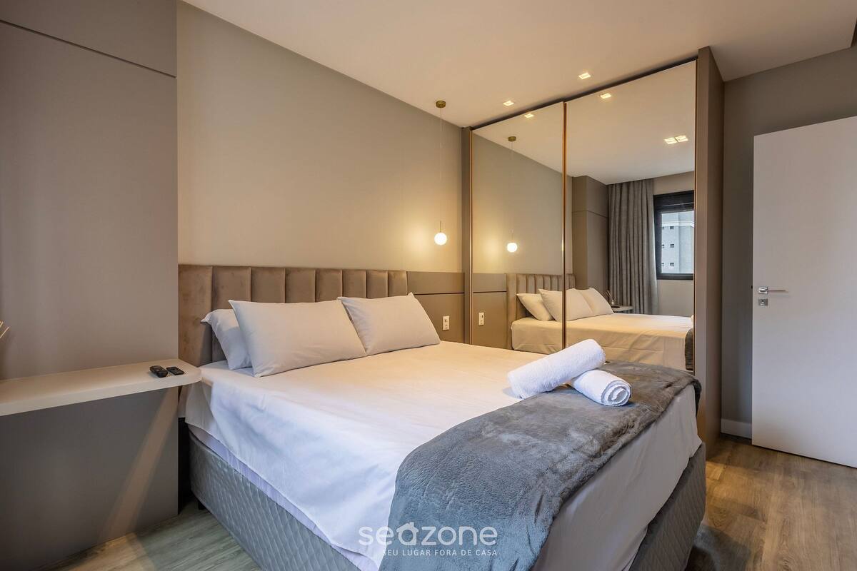 A modern hotel room with a large bed, a bedside table, a mirror, and a view of the outside.