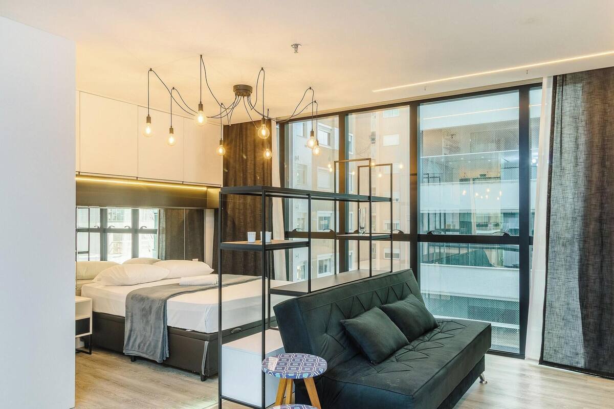 A modern hotel room with a bed, a sofa, a small table, and a large window offering a view of the city.