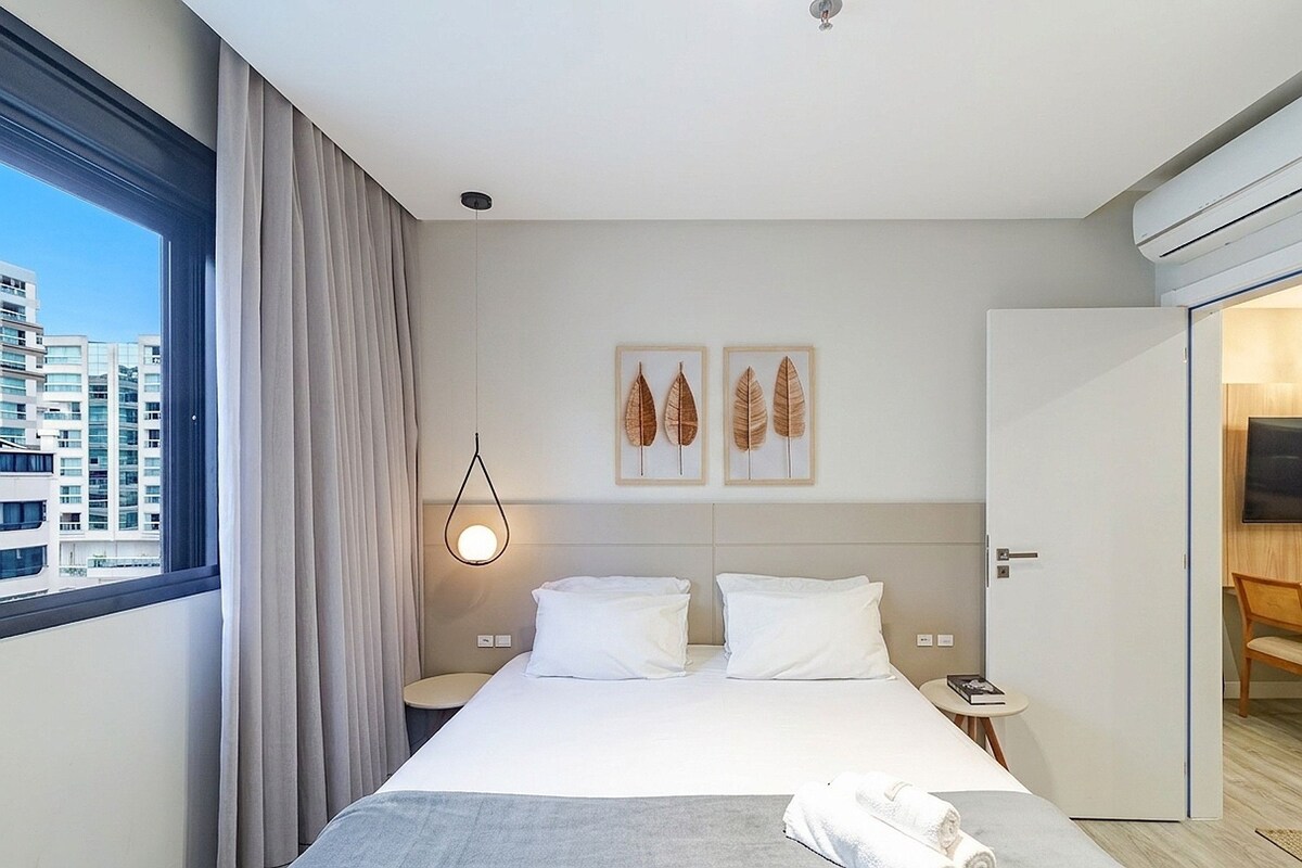 A hotel room with a bed, a bedside table, a nightstand, a window with curtains, a wall-mounted light, and a door leading to another room.