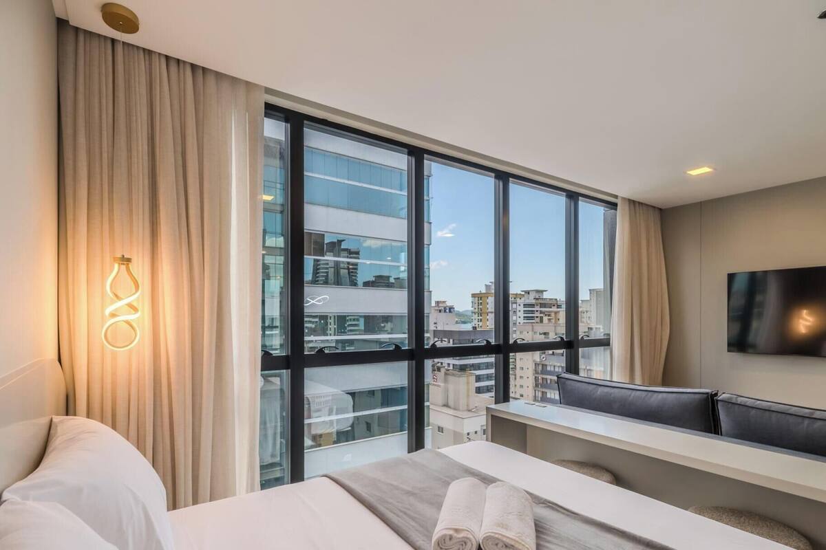 A modern hotel room with a large window, a bed, a desk, and a TV.