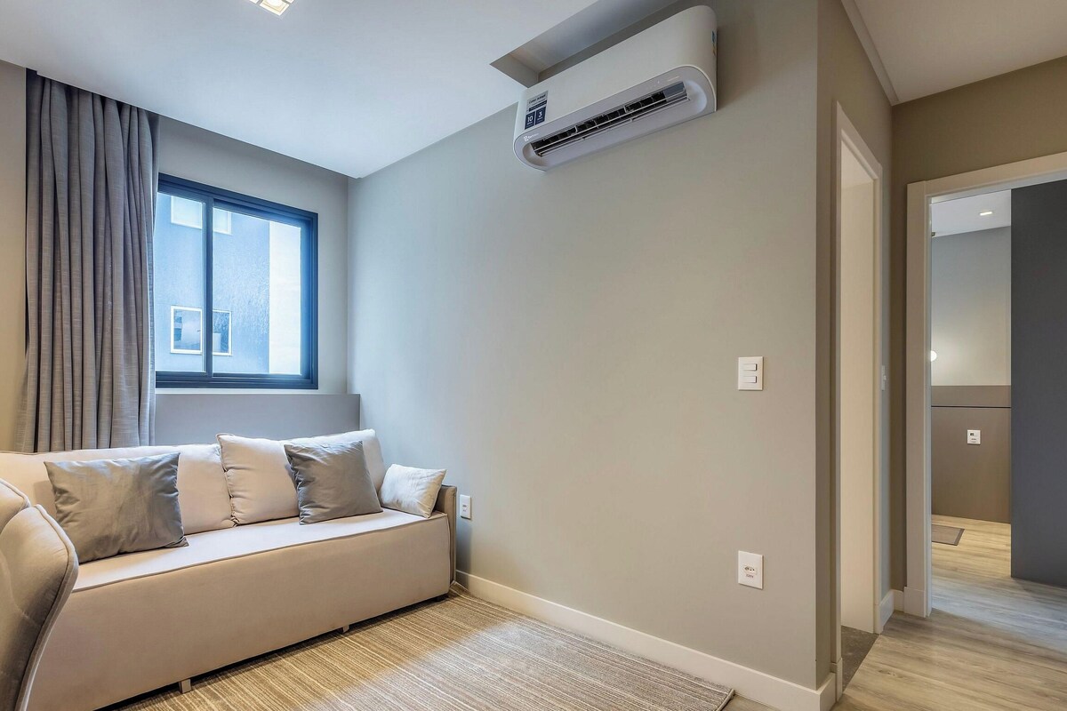 A modern living room with a sofa, a window with curtains, an air conditioning unit, and a hallway leading to another room.