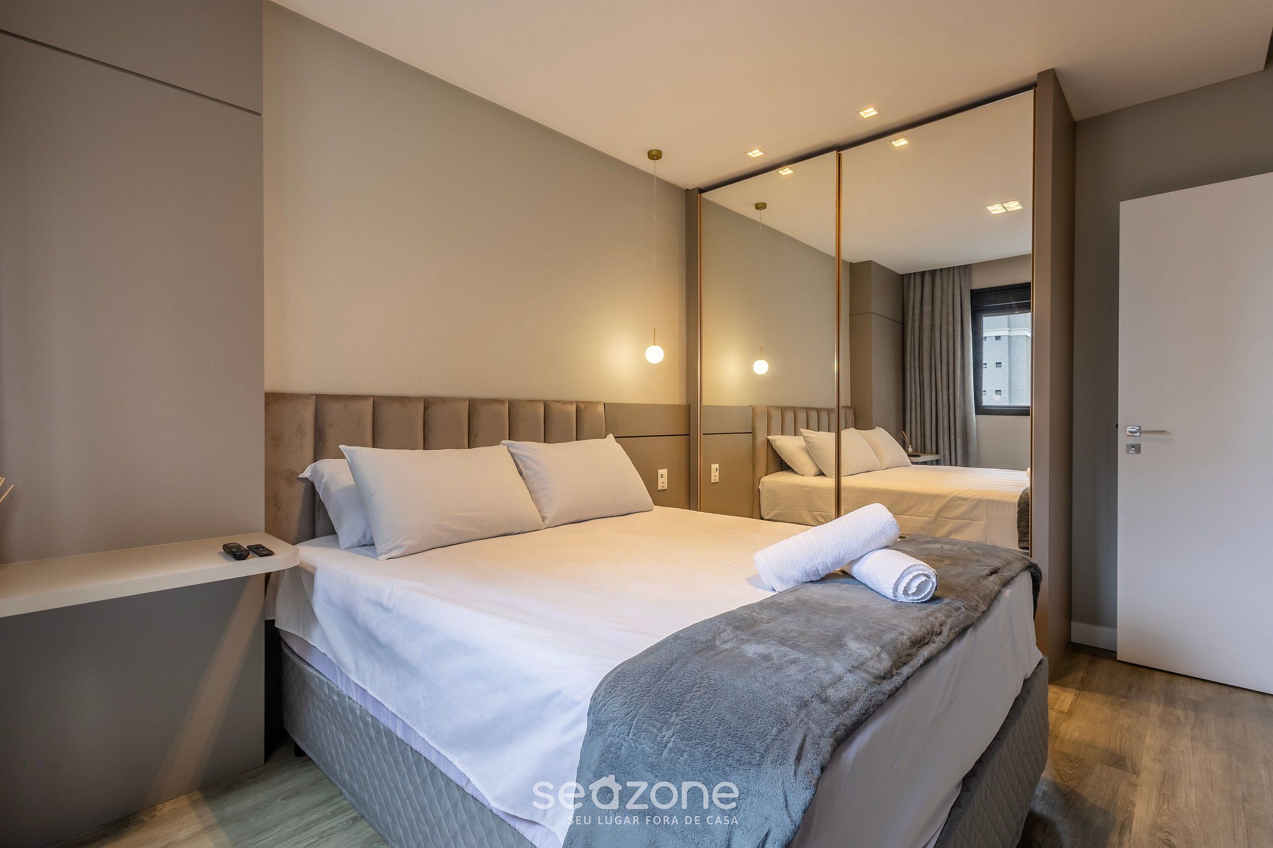 A modern hotel room with a large bed, a bedside table, a mirror, and a view of the outside.