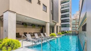 Una piscina con sillas de descanso y un edificio de varios pisos y ventanas.