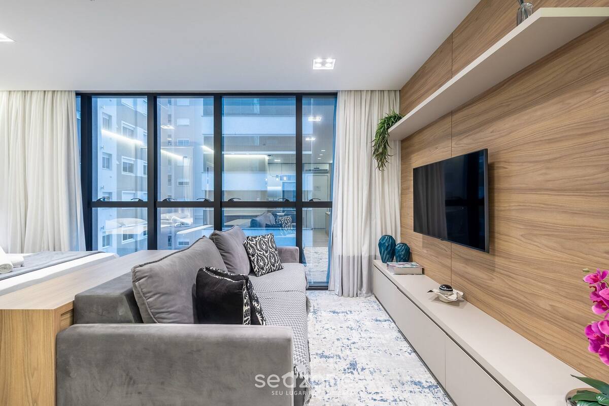 A modern living room with a grey sofa, a flat-screen TV mounted on a wooden wall, and a large window offering a view of the city.