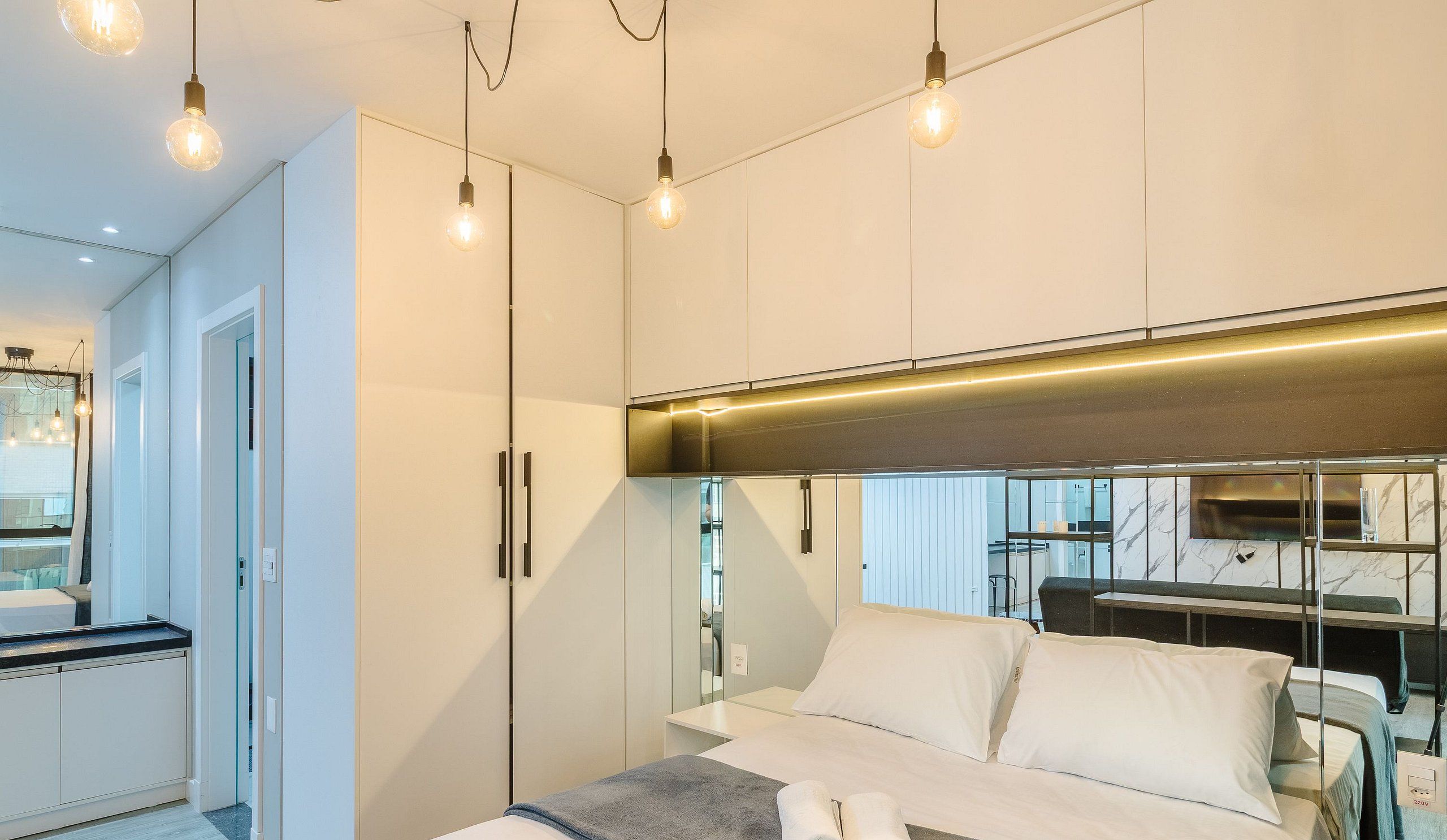 Quarto moderno com cama, mesas de cabeceira e um espelho. Há luminárias no teto e uma área de cozinha planejada, com bancada e armários.
