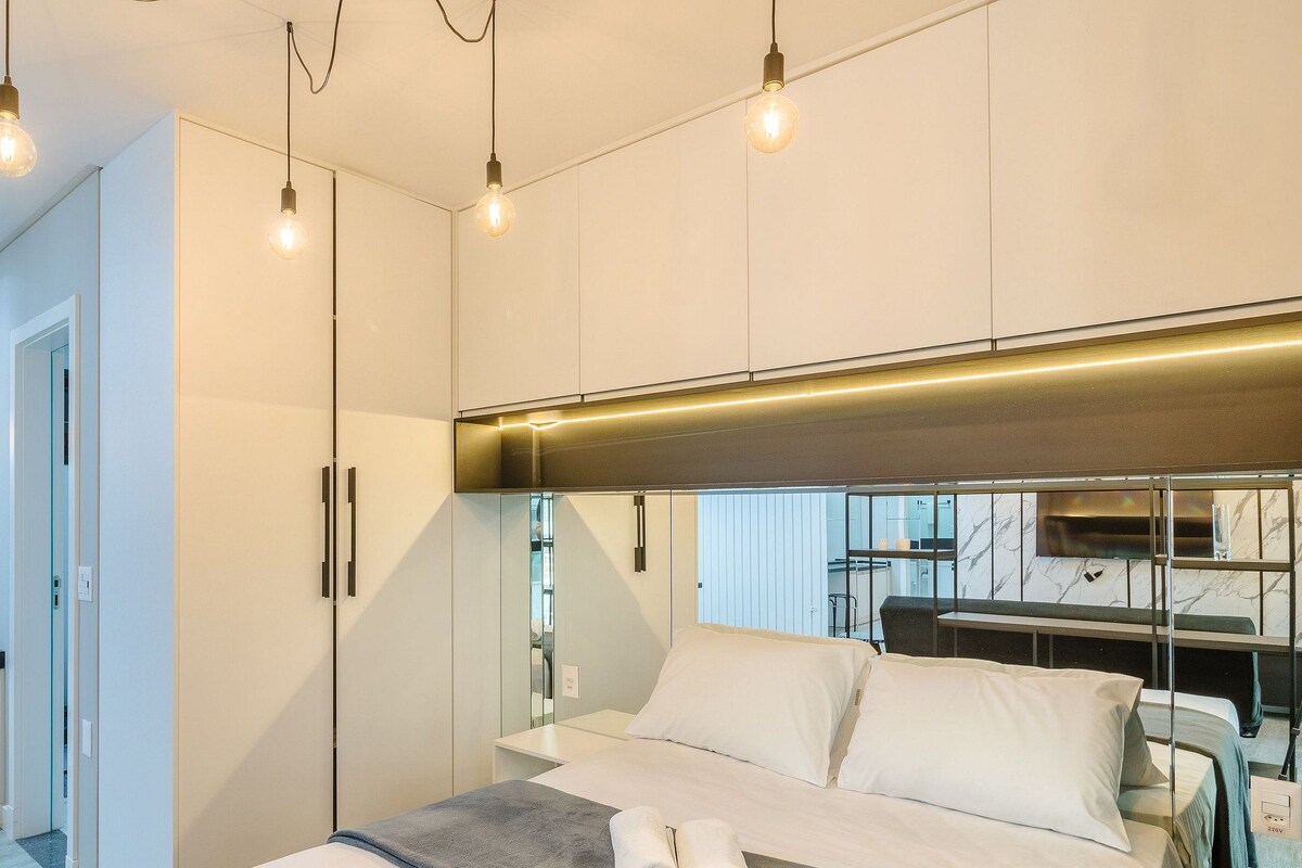 A modern bedroom with a bed, bedside tables, and a mirror. There are light fixtures on the ceiling and a built-in kitchen area with a countertop and cabinets.