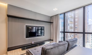 A modern living room with a flat-screen TV mounted on a wall with vertical blinds, a sofa, and a large window offering a city view. - IFT - Ed. Infinity Tower Flat 350m From Meia Praia (Itapema)