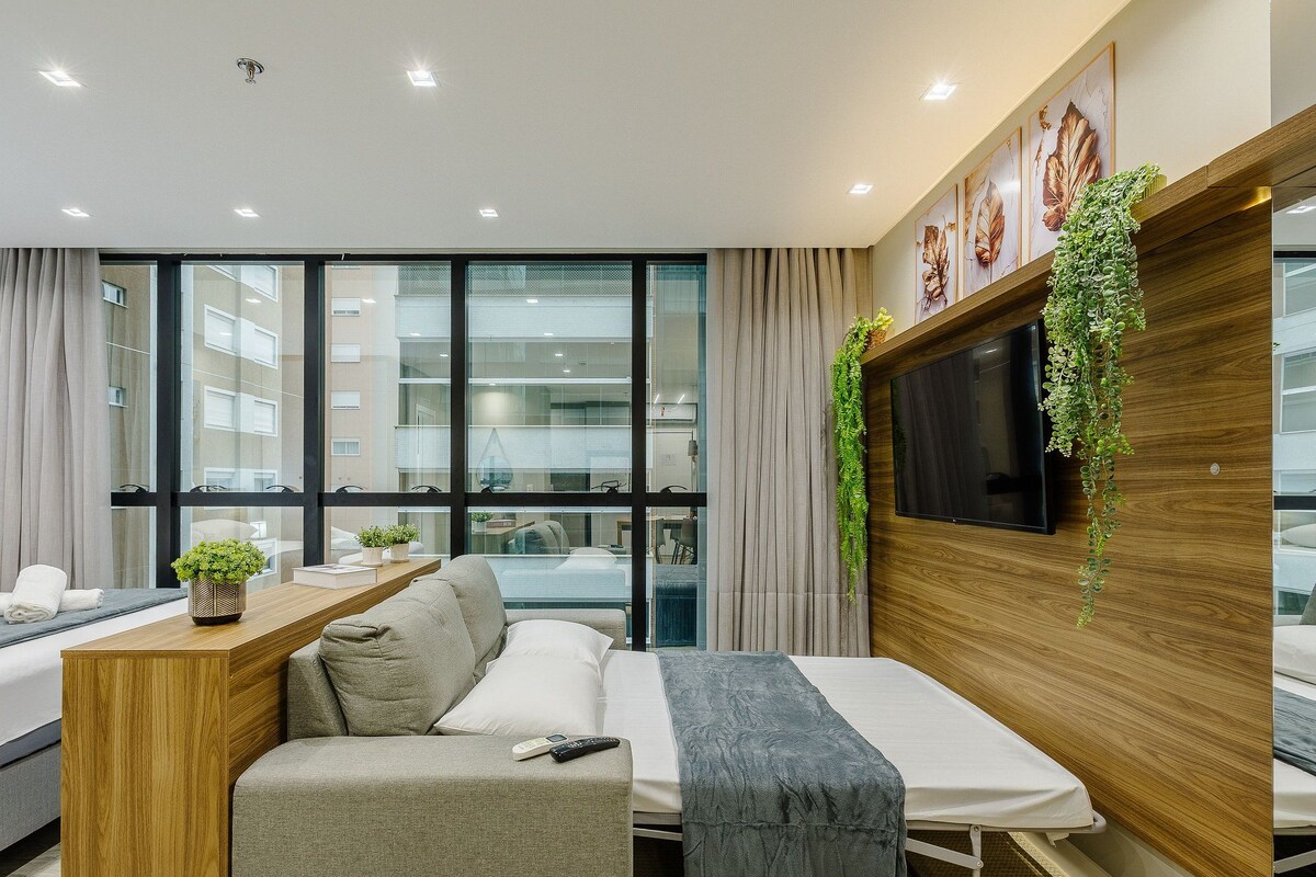A modern hotel room with a sofa bed, a small wooden table with a plant, a TV mounted on a wooden panel, and large windows offering a city view.