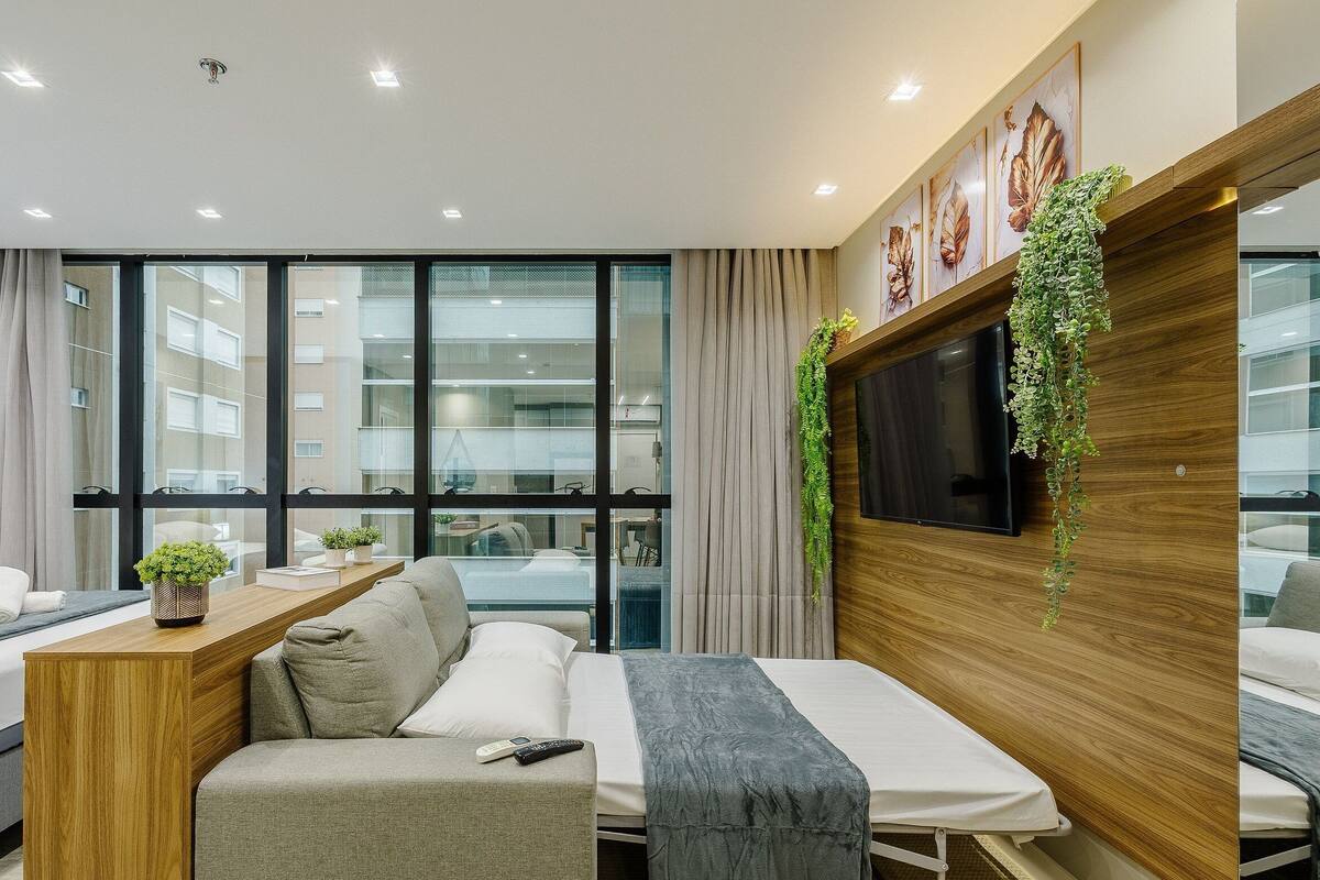 A modern hotel room with a sofa bed, a small wooden table with a plant, a TV mounted on a wooden panel, and large windows offering a city view.