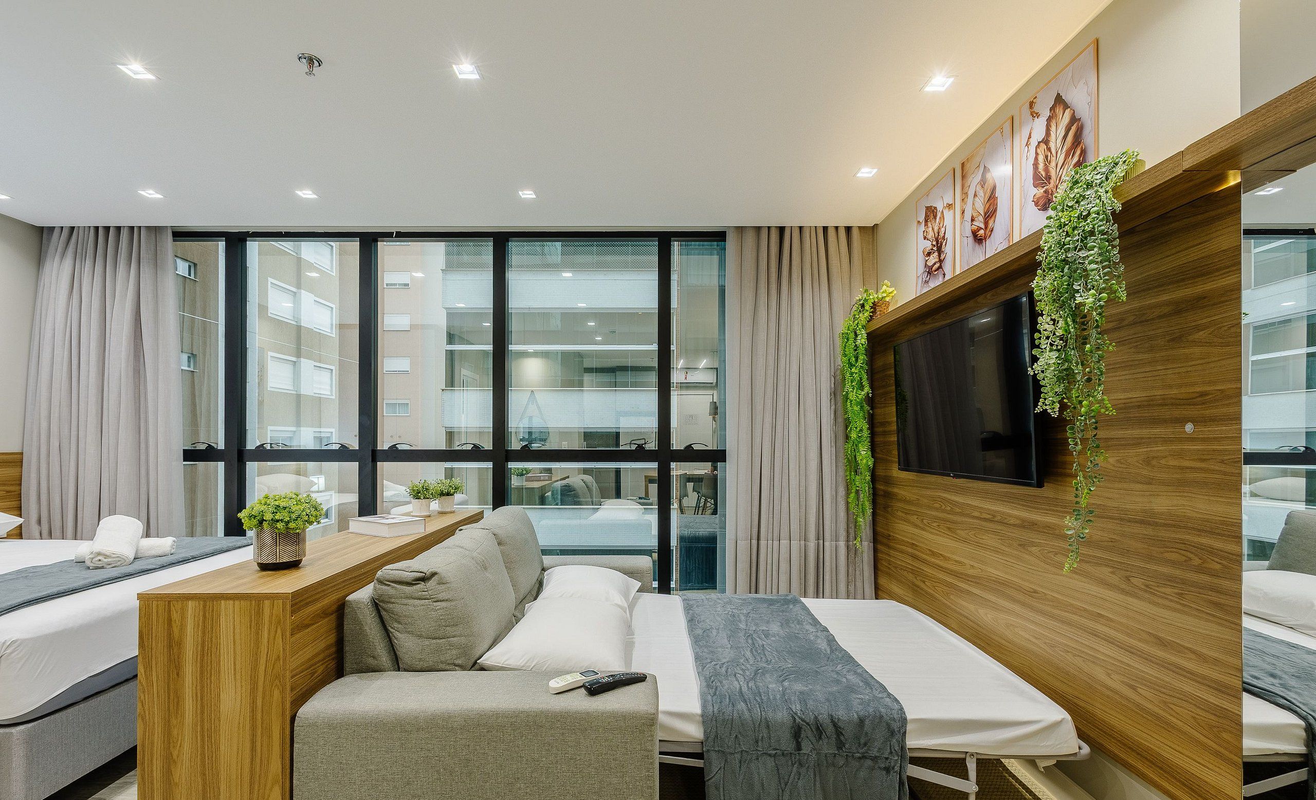 Una habitación de hotel moderna con sofá cama, una pequeña mesa de madera con una planta, un televisor montado en un panel de madera y grandes ventanas con vistas a la ciudad.
