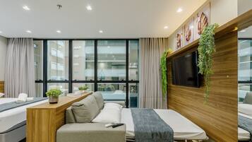 Une chambre d’hôtel moderne équipée d’un canapé-lit, d’une petite table en bois agrémentée d’une plante, d’un téléviseur fixé sur un panneau en bois et de grandes fenêtres offrant une vue sur la ville.