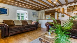Una sala de estar acogedora con un sofá de cuero marrón oscuro, vigas de madera en el techo, una mesa de centro de madera y una escalera que conduce a una zona de altillo.
