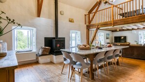 Un interior acogedor y rústico con una mesa de madera central, sillas, una chimenea y una escalera que sube a un nivel superior.
