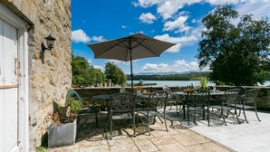 Un patio al aire libre con una mesa y sillas, un paraguas grande y vistas a un lago y montañas.