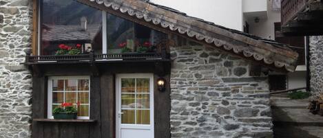 A stone building with a wooden door and windows, a balcony with flowers, and a stone staircase.