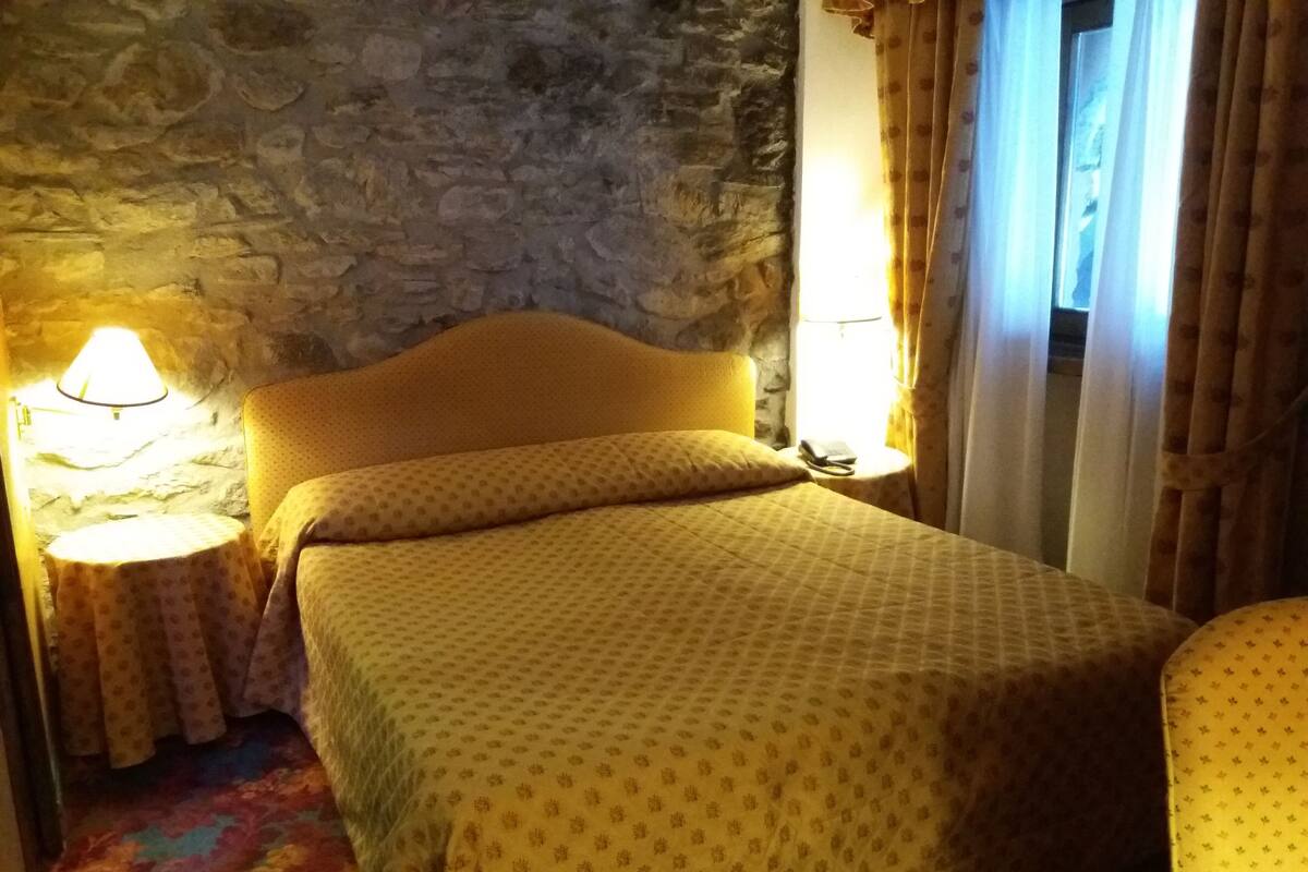 Una habitación con pared de piedra, cama con cabecera amarilla, mesita de noche con lámpara y ventana con cortinas.